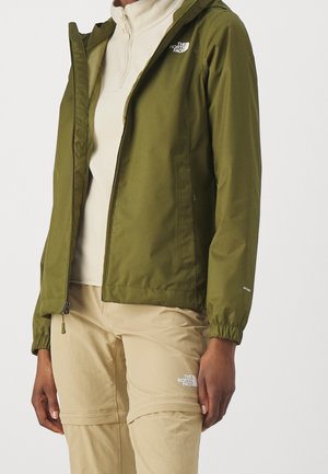 Person trägt olivgrüne Jacke mit Reißverschluss und beige wandelbare Hosen mit Markenlogos, steht vor einfarbigem Hintergrund.