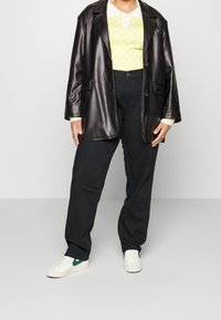 Zwarte neplederen blazer, gele geblokte sweater, zwarte broek, witte sneakers met groene accenten. Casual, moderne outfit met gelaagde texturen.