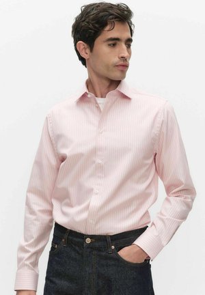 Mand iført en lyserød, stribet skjorte med knapper og lange ærmer samt mørkeblå jeans, kigger mod venstre med den ene hånd i lommen.