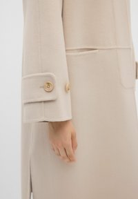 Cappotto in lana beige con un design strutturato, polsini con bottoni chiari e spacco laterale per facilità di movimento.