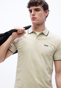 Jeune homme portant un polo beige avec le logo "BOSS", tenant une veste noire sur son épaule sur un fond blanc.
