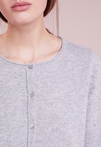 Nahaufnahme einer Person, die eine hellgraue, knöpfbare Strickjacke mit Rundhalsausschnitt vor einem rosa Hintergrund trägt.