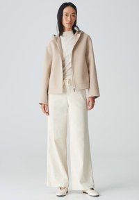 Beige ritsjack met capuchon, van lichtgewicht stof; gecombineerd met crèmekleurige wijde broek en lichte schoenen; gladde textuur en ontspannen pasvorm.