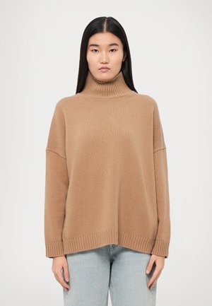 Maglione beige a collo alto in maglia a costine, con spalle scese, una vestibilità rilassata e un orlo con un sottile bordo a costine. Indossato con jeans chiari.