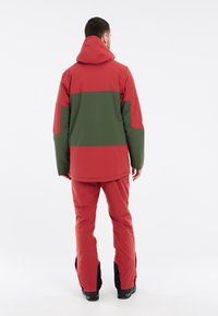 Rote und grüne wasserdichte Jacke mit Kapuze und passenden Schneehosen. Flachnähte, lange Ärmel und Reißverschlüsse an den Knöcheln der Hose.
