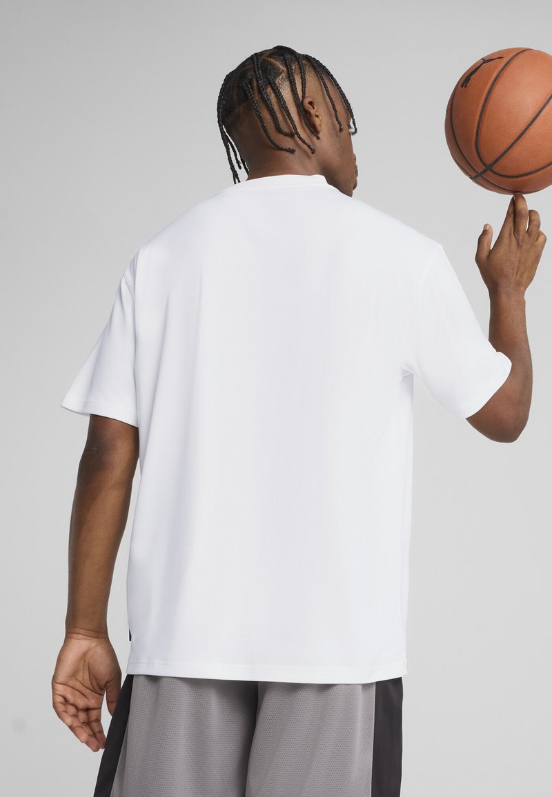 Hombre con cabello trenzado, vistiendo una camisa blanca y pantalones cortos grises, haciendo girar un baloncesto en su dedo, de espaldas, sobre un fondo liso.