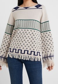 Pull crème tricoté avec divers motifs géométriques en vert, bleu marine et gris. Présente des franges à l'ourlet et des manches larges.