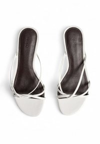 Vita lädersandaler med tunna, korsande remmar över en mörkbrun innersula. Platt sula, öppen baksida och minimal design.