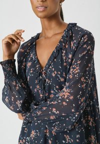 Vrouw in een marineblauwe bloemenjurk met ruches bij de V-hals en smockmanchetten, met een hand tegen haar gezicht voor een effen achtergrond.