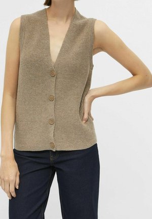Personne portant un gilet en tricot beige sans manches boutonné, associé à un jean bleu foncé, main posée sur la hanche sur un fond uni.
