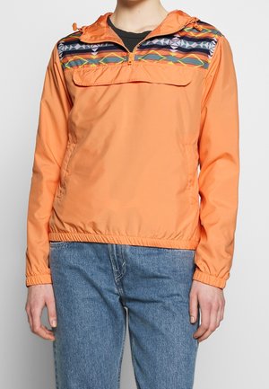 Osoba ubrana w pomarańczową kurtkę z kolorowym geometrycznym wzorem na klatce piersiowej, w połączeniu z niebieskimi dżinsami.