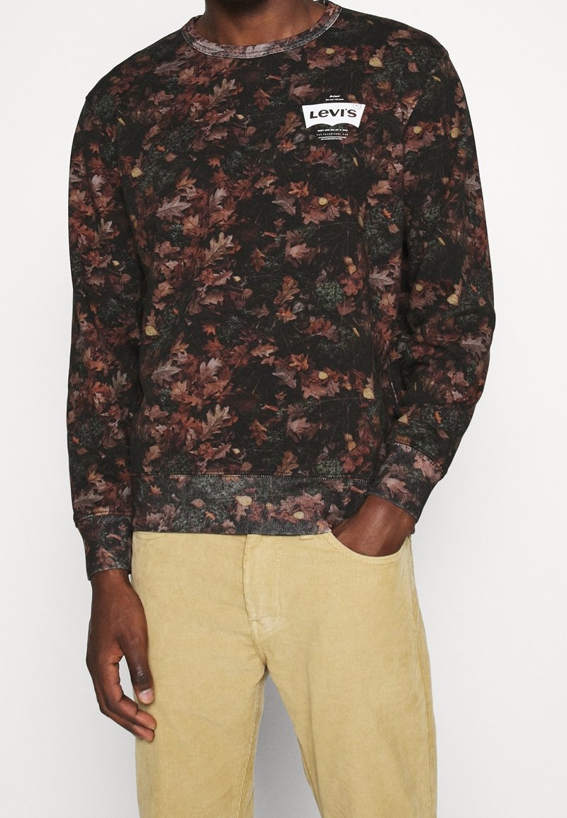 Homme portant une chemise à manches longues foncée avec un motif de feuilles marron et vertes et un petit patch logo Levi's blanc, associée à un pantalon beige clair.