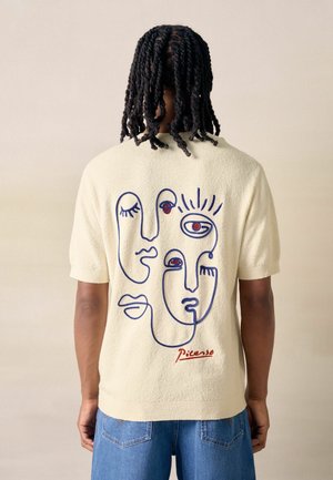 Man with braided hair wearing cream short-sleeve shirt featuring blue continuous line faces and red "Picasso" signature on back.