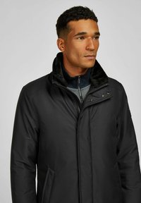 Zwarte gewatteerde jas met een getextureerde buitenkant, voorzien van een nepbontkraag, gesloten met drukknopen en een rits aan de voorkant. Inclusief zijzakken.