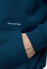 Χέρι στην τσέπη ενός σκούρου πετρόλ φούτερ Hollister που εμφανίζει ριμπ μανσέτα και κεντημένο όνομα επωνυμίας πάνω από την τσέπη.