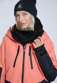 Koralfarvet og sort vandtæt jakke med justerbar hætte, lynlåslommer, forstærkede sømme, parret med et sort strikket tørklæde og hue.