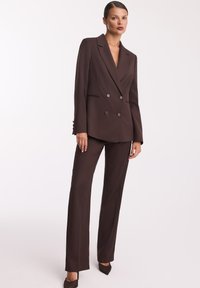 Dwurzędowy brązowy garnitur z gładkiego materiału, z szerokimi klapami, dwoma guzikami oraz prostymi spodniami o czystym wykończeniu.