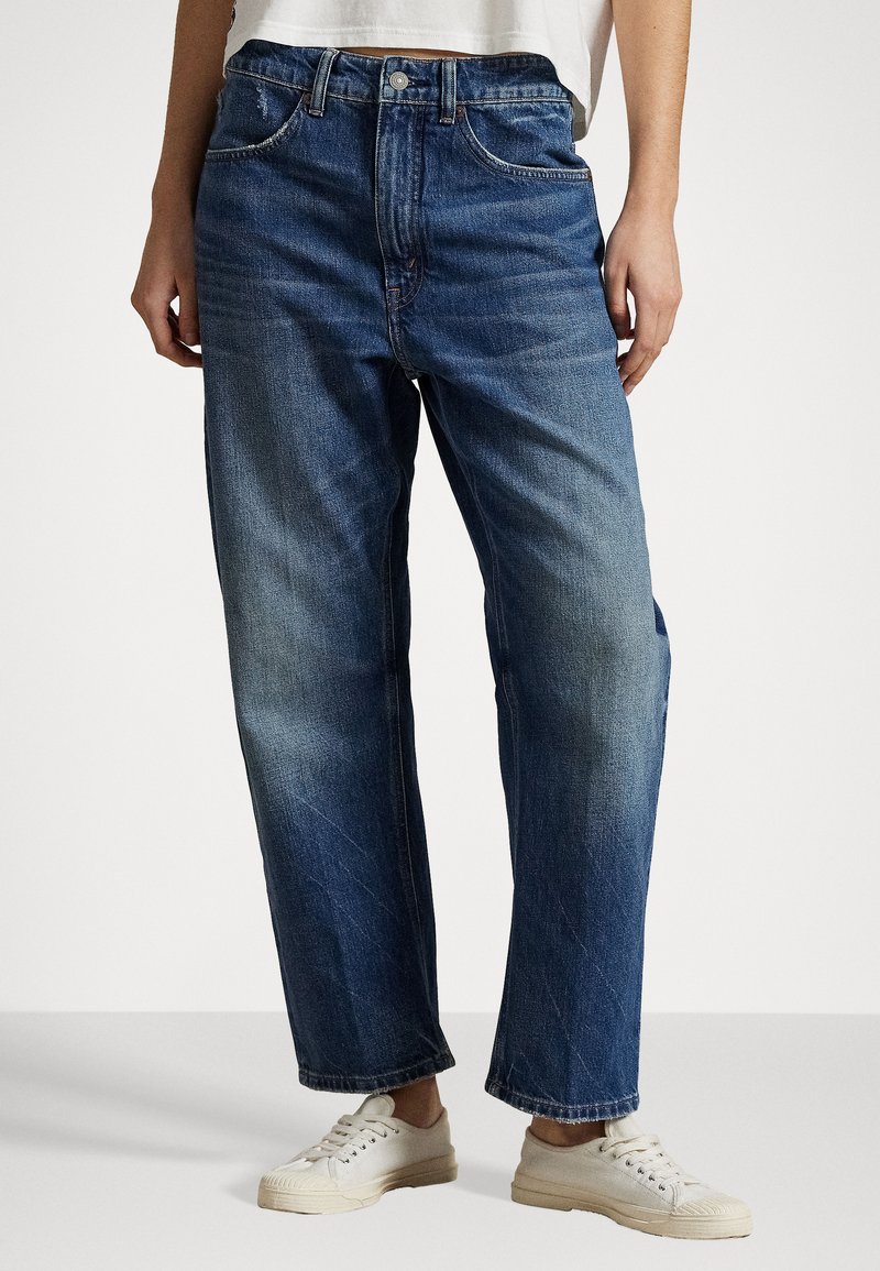 Denim jeans with a relaxed fit, medium blue color, slight fading, five pockets, belt loops, and a single button closure, paired with white sneakers.