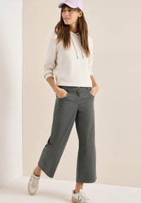 Mujer con cabello largo y castaño, que lleva una sudadera con capucha de color beige claro, pantalones anchos de color gris oscuro, zapatillas blancas y una gorra de béisbol rosa claro, de pie con las manos en los bolsillos.