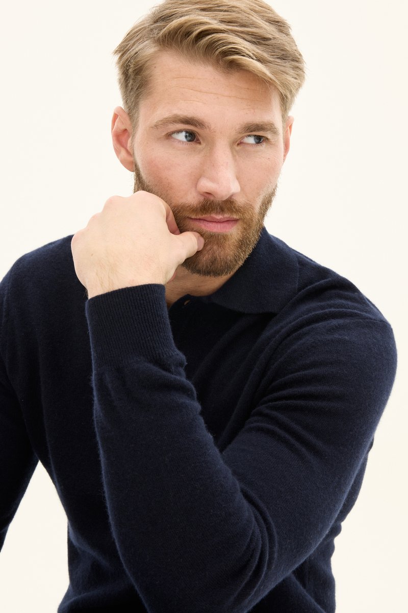 Navyblaues Strickpolo aus leichtem Stoff mit einem Kragen. Die Hand des Models ruht auf dem Kinn und zeigt den Ärmel und die Textur.