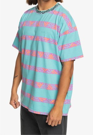 Osoba ubrana w turkusowy T-shirt z różowymi i pomarańczowymi zygzakowatymi paskami oraz czarne spodnie, pokazana od ramion do bioder.