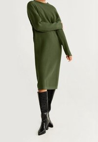Olivgrünes, geripptes Strickkleid mit langen Ärmeln, Rundhalsausschnitt und knielangem Schnitt. Tragen mit glänzenden, schwarzen Stiefeln bis zum Knie.