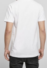 Muž v jednoduchém bílém tričku s krátkým rukávem a černých džínách, zobrazený zezadu na neutrálním světle šedém pozadí.