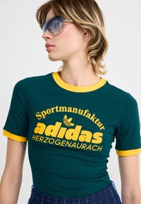 Green cotton t-shirt with yellow ribbed neckline and sleeves. Features large yellow text saying "adidas" and a logo, fitted design.
