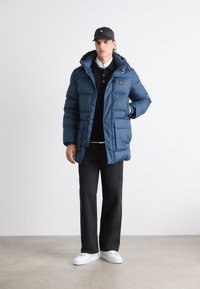 Giacca blu imbottita con cappuccio, dotata di zip frontale, tasche e polsini elastici, abbinata a un polo nera e pantaloni scuri. Sneakers bianche.