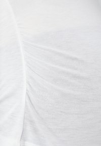 Tela de algodón blanco con una textura suave y un ligero estiramiento. Contiene detalles fruncidos a lo largo de la costura lateral. Diseño simple y limpio.