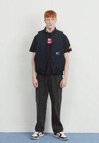 Ναυτικό αμάνικο γιλέκο πάνω από μαύρο t-shirt, με ροζ λογότυπο. Μαύρα παντελόνια σε φαρδιά γραμμή και μαύρα-Λευκά αθλητικά συμπληρώνουν την εμφάνιση.