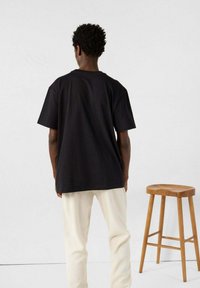 Schwarzes, übergroßes T-Shirt aus Baumwolle mit kurzen Ärmeln und Rundhalsausschnitt, kombiniert mit hellcremefarbenen Hosen und einem Holzstuhl.