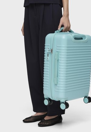 Personne portant un pantalon noir et des chaussures, tenant une valise rigide bleu clair avec quatre roues et une serrure intégrée.