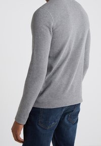 Graues Langarmshirt mit geraffter Struktur, rundem Ausschnitt und schmaler Passform, kombiniert mit blauen Jeans mit Gesäßtaschen.