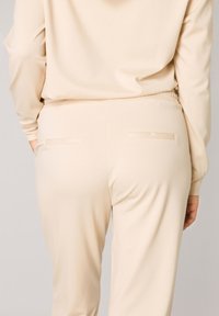 Beige trainingsbroek van zacht materiaal, met een elastische tailleband en twee achterzakken met platte sluitingen. Soepele textuur.