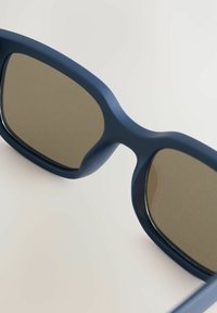 Occhiali da sole con montatura in plastica opaca color navy, forma rettangolare e lenti marrone specchiate. Presentano bordi arrotondati e hardware minimal.