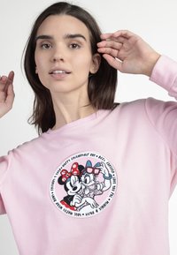 Joven mujer vestida con una sudadera de color rosa claro que presenta a Minnie Mouse y Daisy Duck abrazándose, con un texto temático de amistad alrededor de ellas.