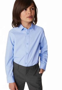 Camicia azzurro chiaro con bottoni, colletto, maniche lunghe e texture liscia, abbinata a pantaloni grigio scuro. Bambino in piedi in modo disinvolto.