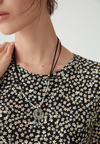 Schwarzes Oberteil mit floralem Muster in kleinen gelben und blauen Blumen, dazu eine silberfarbene Halskette mit einem runden Anhänger und Perlen.