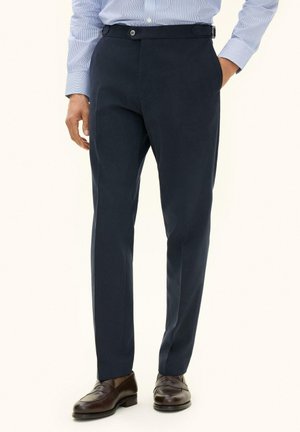 Mann trägt marineblaue formale Hose, hellblaues Hemd und dunkelbraune Lederloafers, steht mit einer Hand in der Tasche.