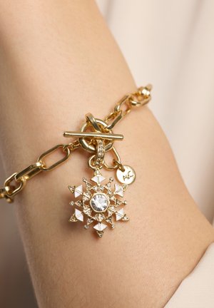 Pulsera de cadena dorada con cierre de barra y un colgante en forma de copo de nieve con esmalte blanco y cristales transparentes en una muñeca.