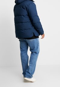 Mørkeblå dunjakke med vandrette quiltede sektioner, hætte og sideslids, kombineret med lyseblå jeans og hvide sneakers.