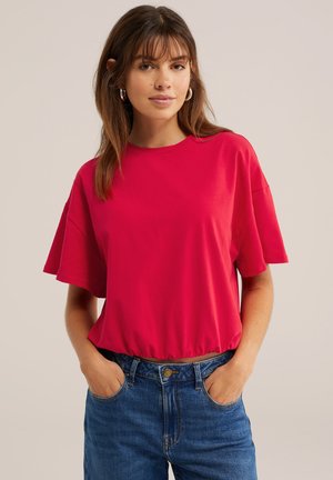 Jeune femme portant un t-shirt ample rouge à manches courtes et un jean bleu, les mains dans les poches, sur un fond uni clair.