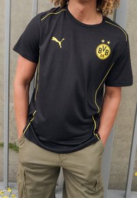 Osoba v černém sportovním tričku Puma se žlutým logem BVB 09 a žlutými lemovkami, v kombinaci s béžovými cargo kalhotami, stojící s rukama v kapsách.