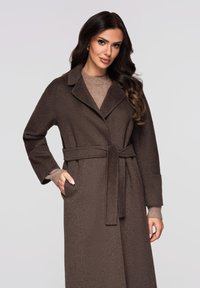 Brun frakke i uldblanding med bælte, hakket krave, lange ærmer og sidelommer. Blød tekstur; lagdelt over en lysebrun top.