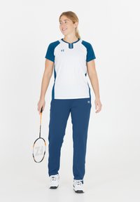 Kvinna i blå och vit sportklädsel som håller en badmintonracket, står och ler, mot en neutral vit bakgrund.