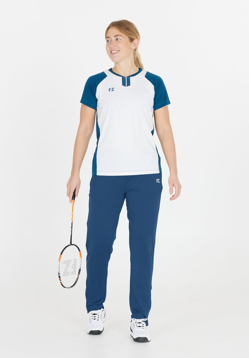 Kvinna i blå och vit sportklädsel som håller en badmintonracket, står och ler, mot en neutral vit bakgrund.