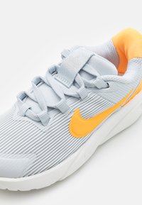 Chaussure de sport gris clair avec des nervures verticales, accent Swoosh orange et tige en maille texturée. Présente des lacets plats et un col rembourré.
