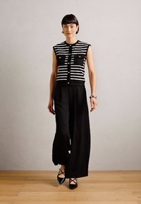 Haut sans manches à rayures noires et blanches avec deux poches poitrine, associé à un pantalon large noir et des chaussures noires à bouts pointus. Texture de tissu lisse.