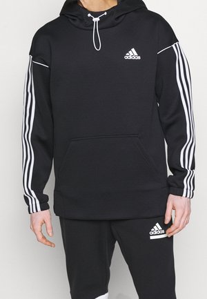 Hombre con sudadera con capucha Adidas negra y pantalones a juego con rayas blancas en las mangas y logotipos en el pecho y el muslo.